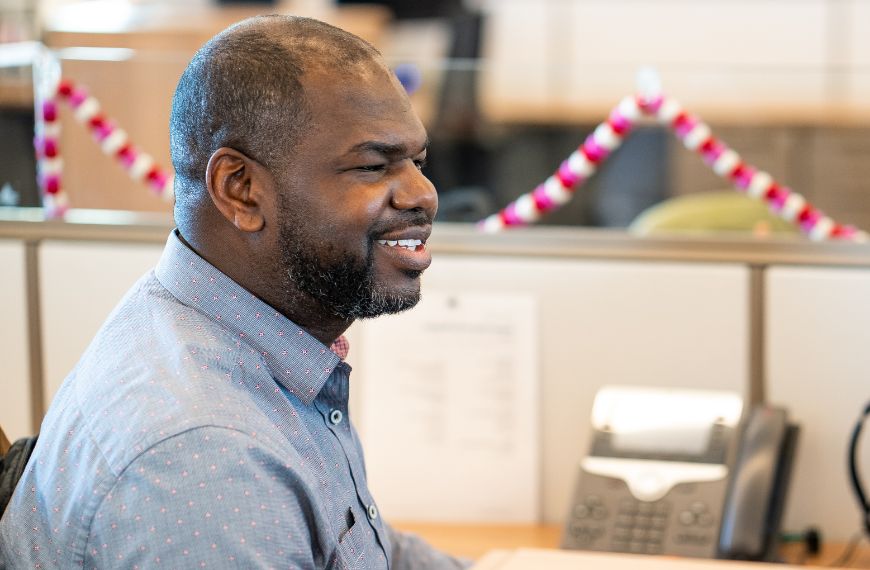 A male Heartland employee is sitting in a cubicle and smiling at his computer 