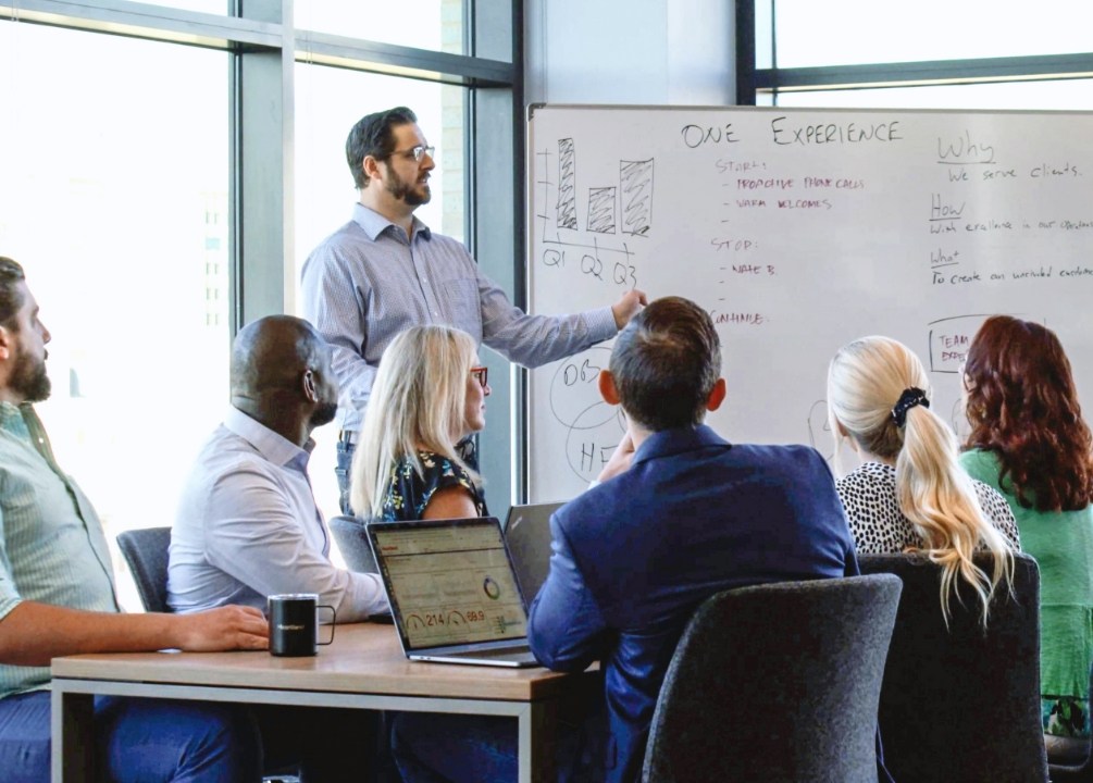 A man points at a whiteboard while giving a presentation to five other Heartland employees. 