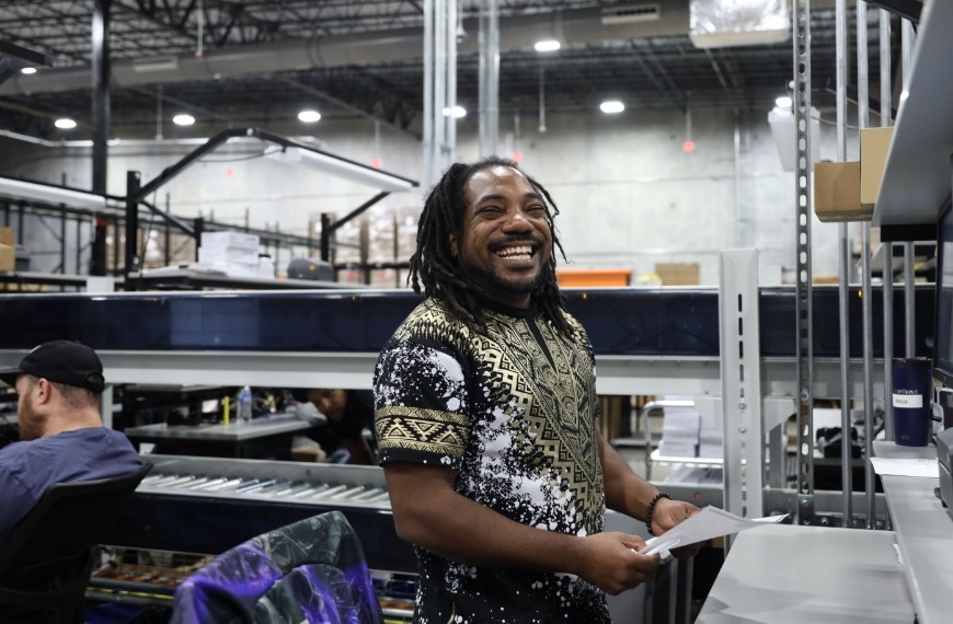 A man with dreadlocks smiles as he holds a paper in a warehouse setting.