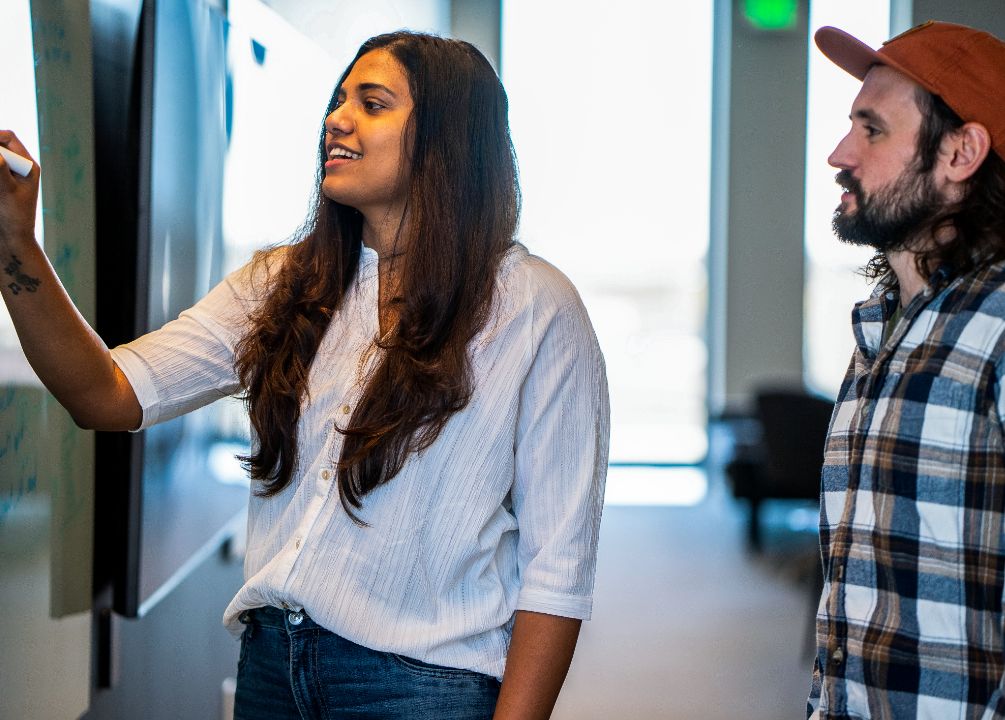 A female Heartland employee with long hair, white blouse and jeans writes code on a whiteboard while a male colleague stands reading her writing.