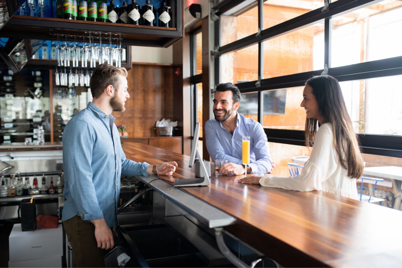 Two customers at a bar top with drinks talking to a man behind the counter. 