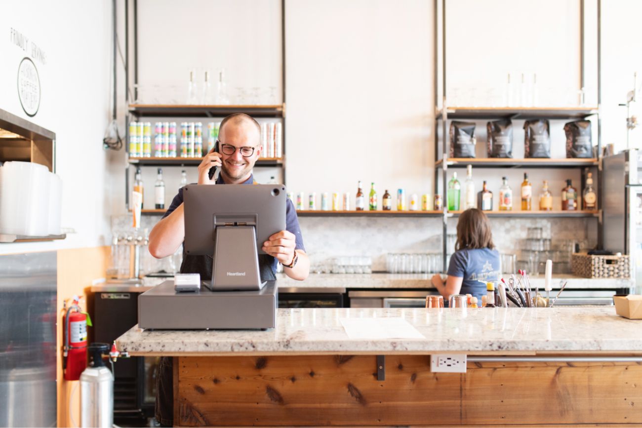A male employee standing at the POS system while talking on the phone and a woman employee making coffee in the background.
