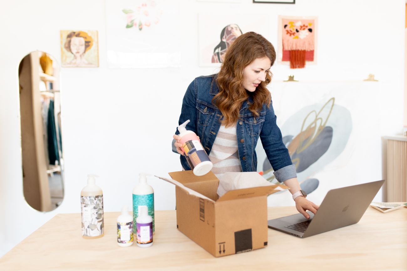 A woman packing bottled products into a bag for shipping while looking at a computer.
