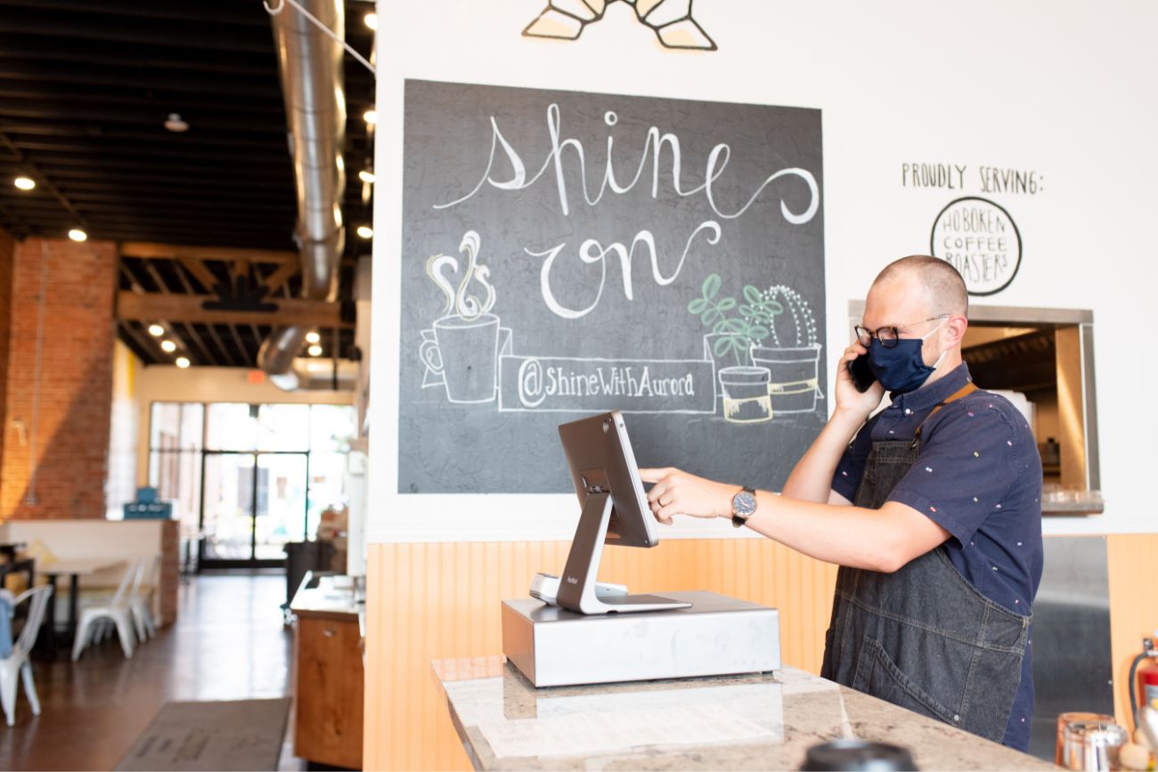 A man working at a coffee shop looking at a screen while talking on the phone.