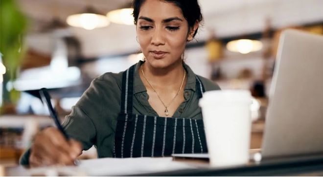 Small business owner writing, laptop open for a productive business day.