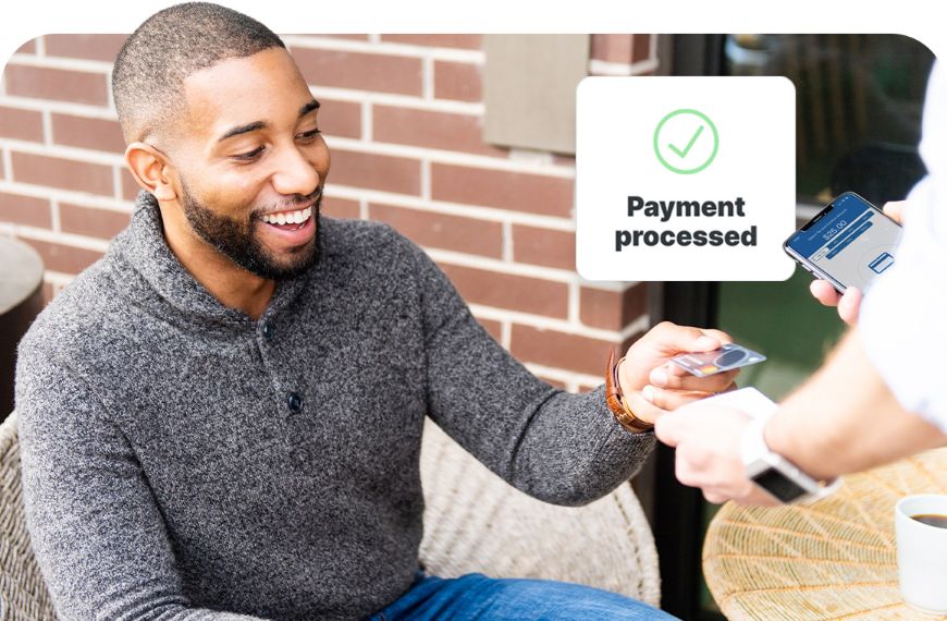 A graphic representation of a payment being processed with a checkmark appears between a smiling man at a table as he taps his card against a payment device and a man holding the payment device and a phone.