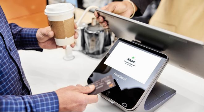 A closeup of a person holding a to-go cup of coffee in one hand and using the other hand to tap a card against a mobile point of sale device screen.