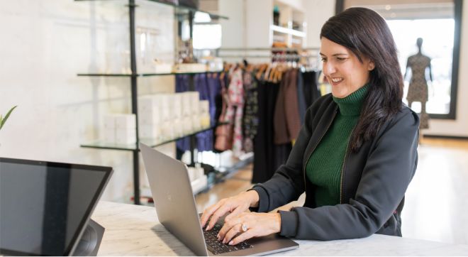A smiling woman in a retail setting sits at a countertop and types on her laptop computer.