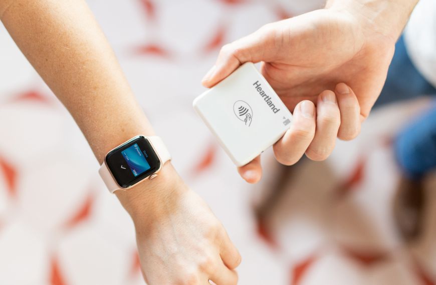 A closeup of a persons wrist and smartwatch next to another hand holding a contactless payment device