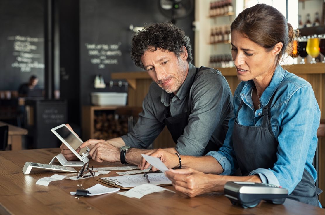A male and female employee sitting in a restaurant with a tablet reconciling multiple receipts