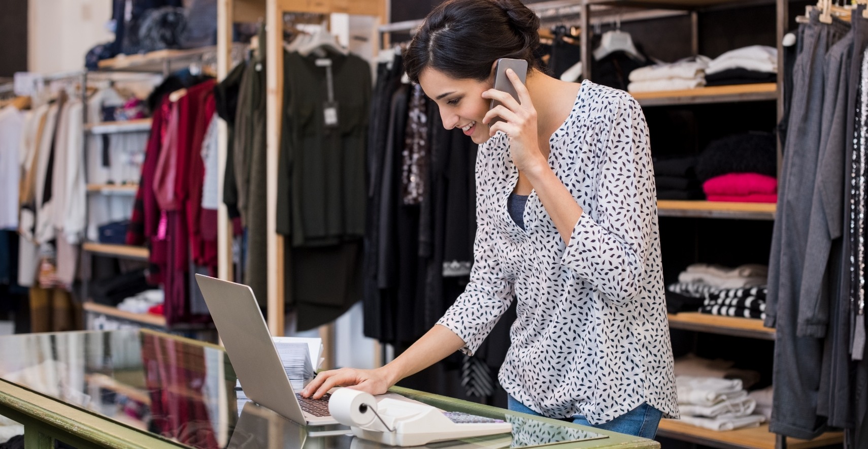 A woman in a retail store talks on the phone while looking at her laptop.