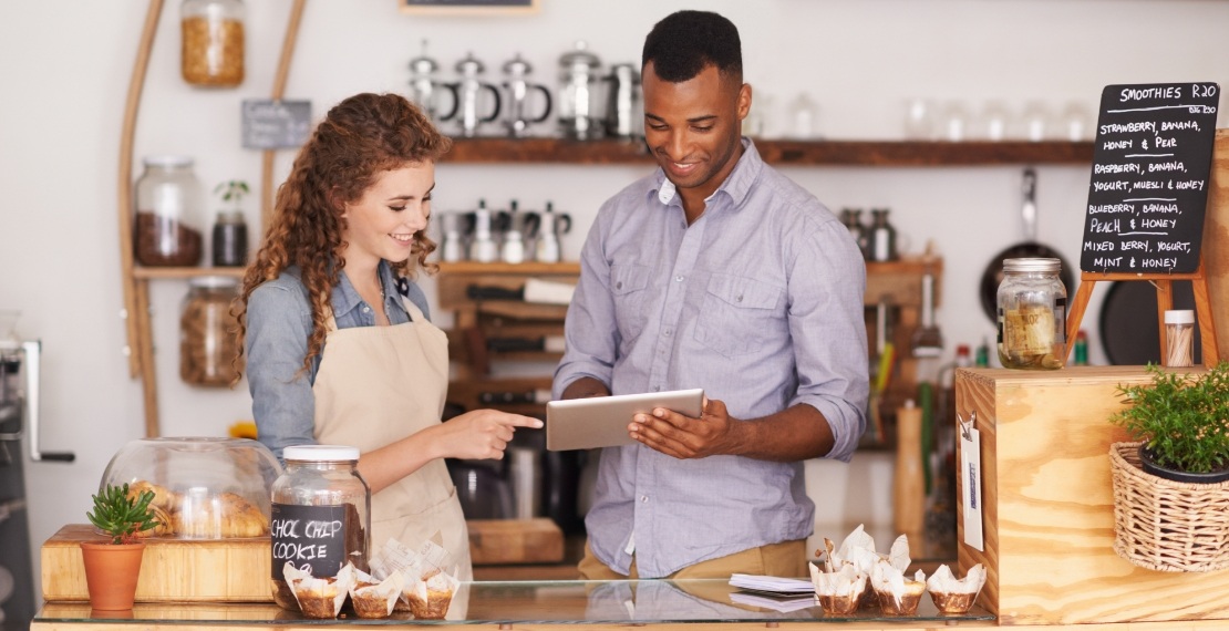 Two employees in a coffee shop talk while using a tablet.