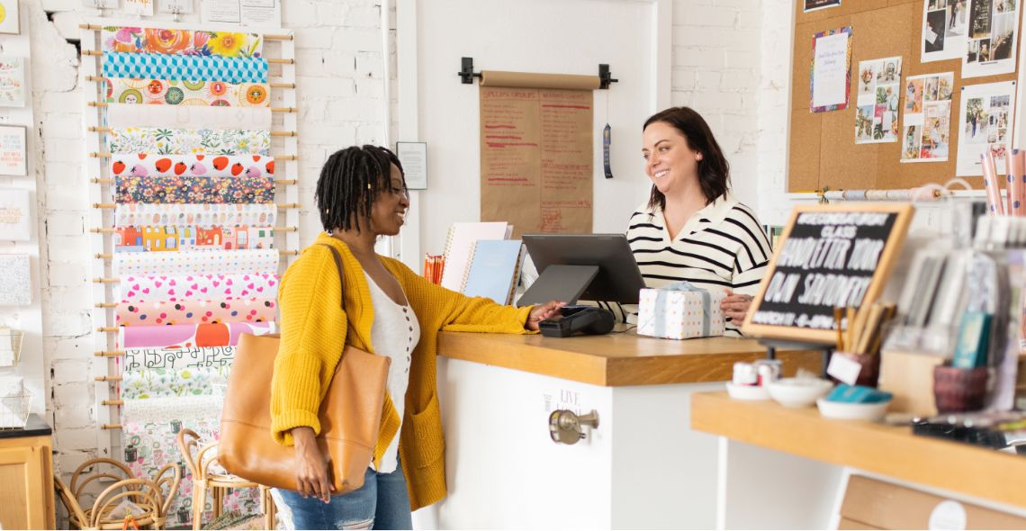 A female customer pays at the point of sale terminal of a retail store while the female retail worker stands behind the counter smiling.