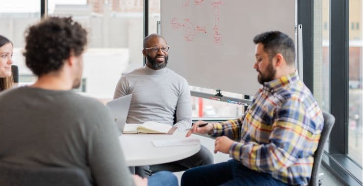 A group of coworkers in an office sit at a table and chat.