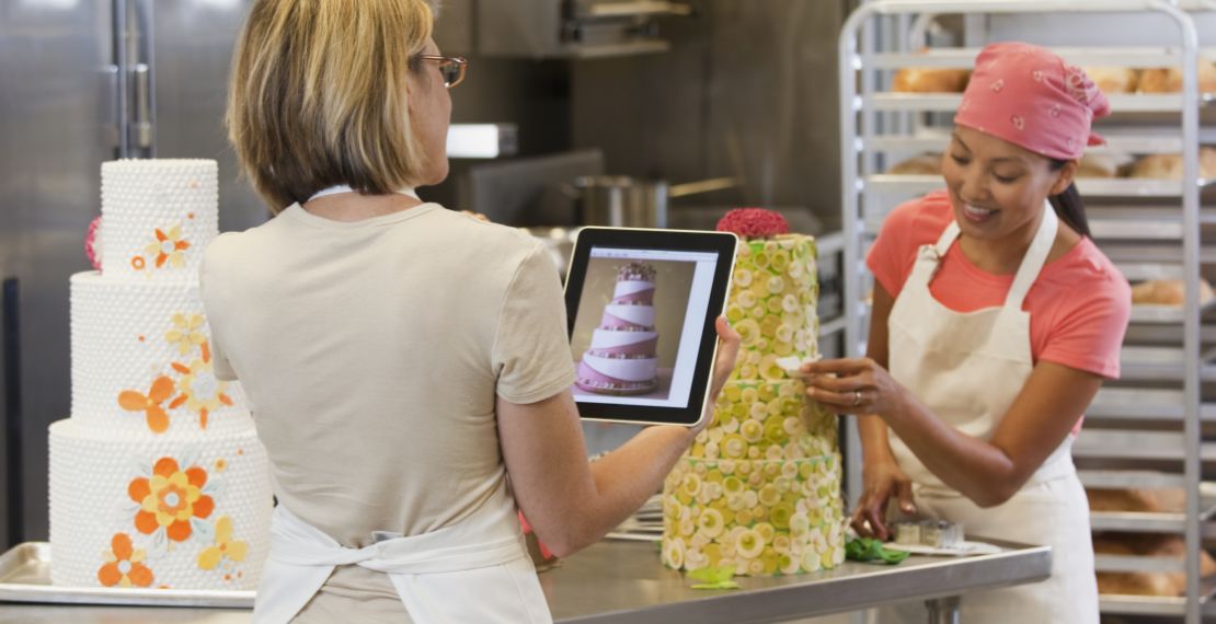 Baker decorates a tiered, yellow cake in a kitchen while consulting with someone about cake design.