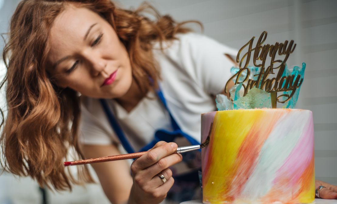 Woman in blue apron decorates a multi-colored, striped birthday cake.