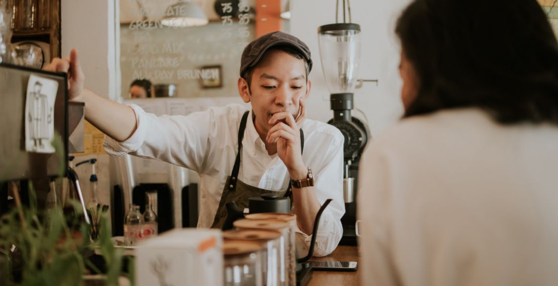 Barista working in a cafe looks contemplative while having a conversation with someone.