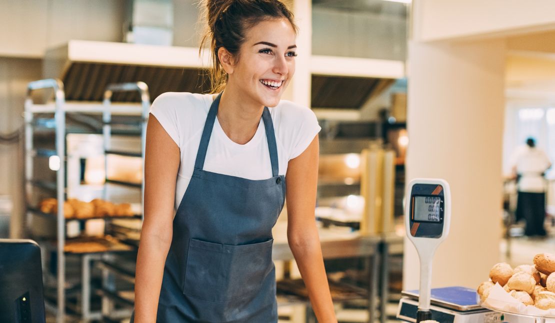 Person standing in a shop with baked goods smiles across the counter.