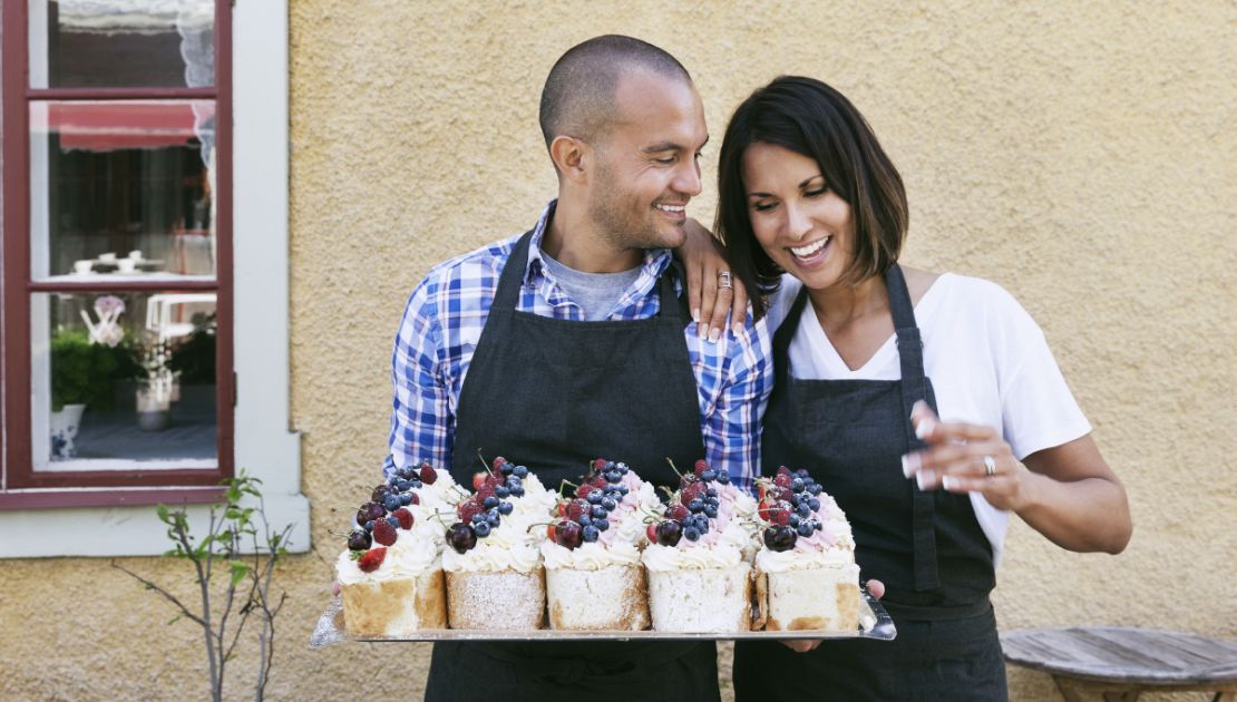 Two people in aprons stand close together and hold a tray of desserts while smiling.