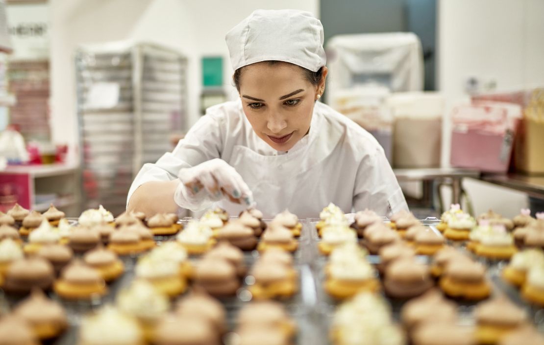 Baker in white hat and coat analyzes trays of desserts sitting on cooling racks.