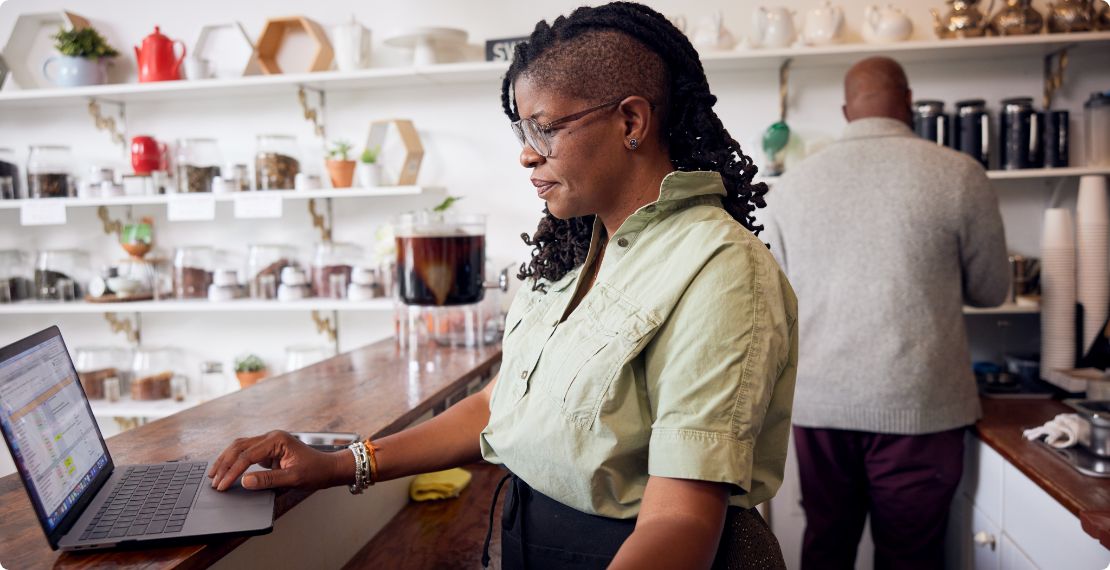 A retail shop owner stands at a counter, scrolling on a laptop.