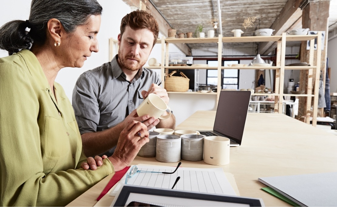 Two people at a workshop table look at ceramic cups. A notepad and laptop sit nearby.
