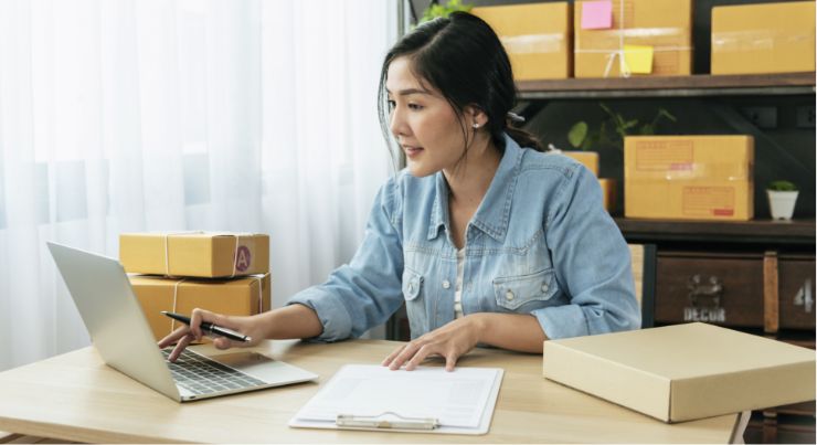 A woman with a pen in hand looks at an open laptop and is sitting at a desk surrounded by packages