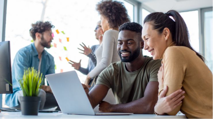 A man and woman look with smiles at an open laptop together while another man and two women have a separate conversation behind them