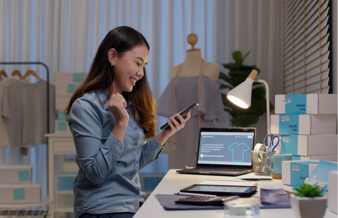 Woman sitting at a desk in the back office of a clothing store, looking at her phone