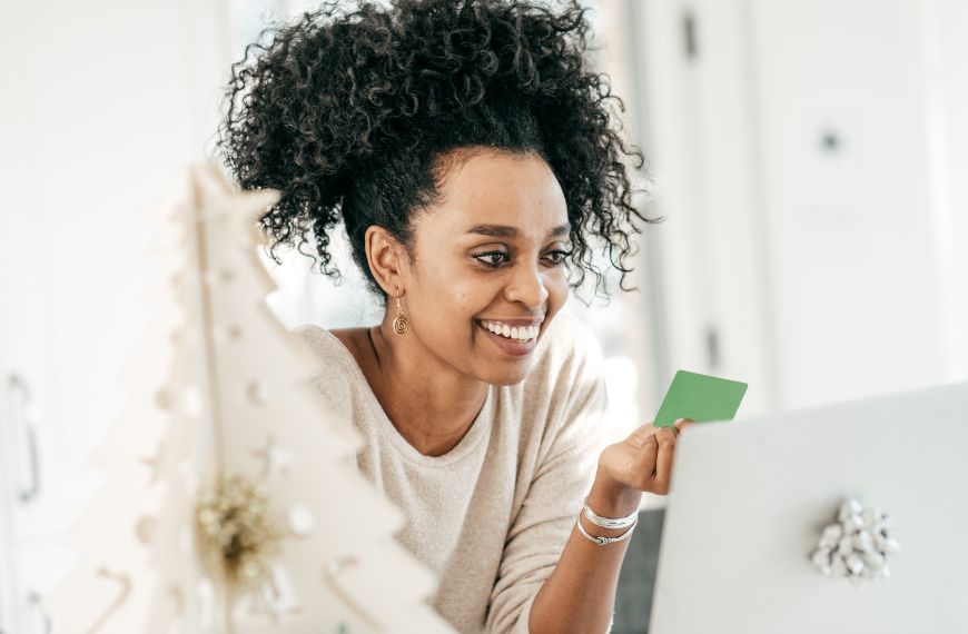 Woman sitting behind white holiday decor, smiling at her laptop and holding a green gift card
