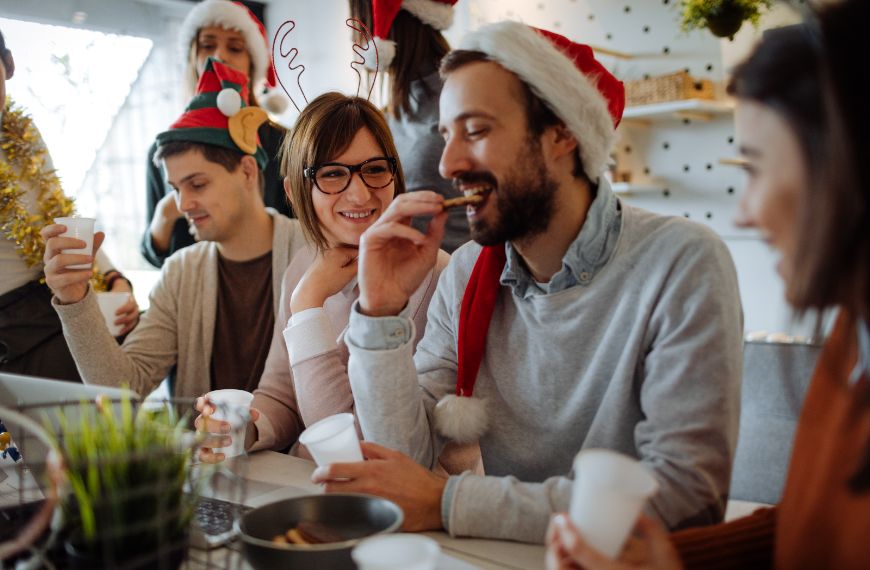 Group of people in Santa and elf hats, smiling and eating around a holiday table