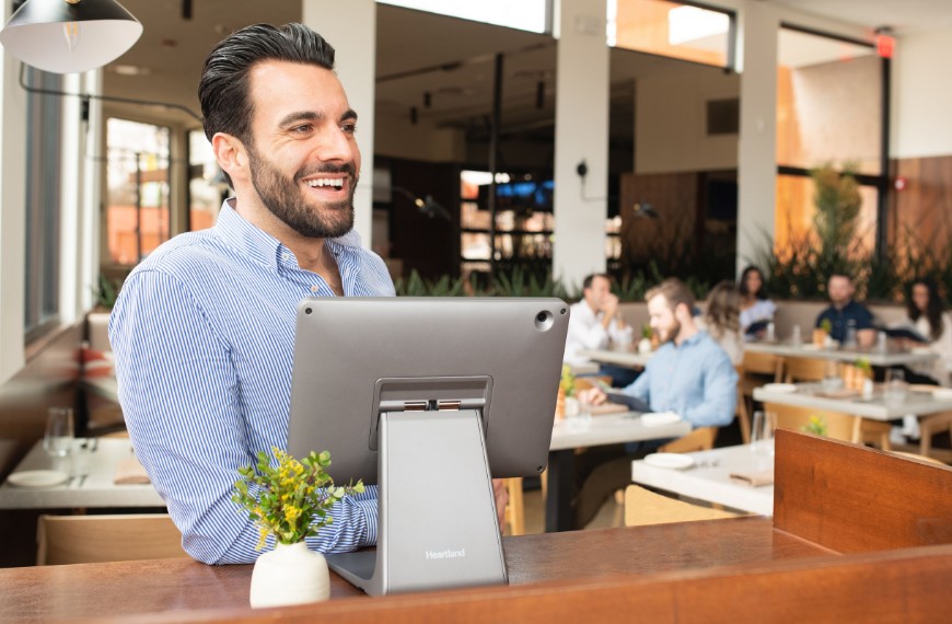 Restaurant host wearing a blue dress shirt leans on his stand, smiling over a Heartland restaurant point of sale system