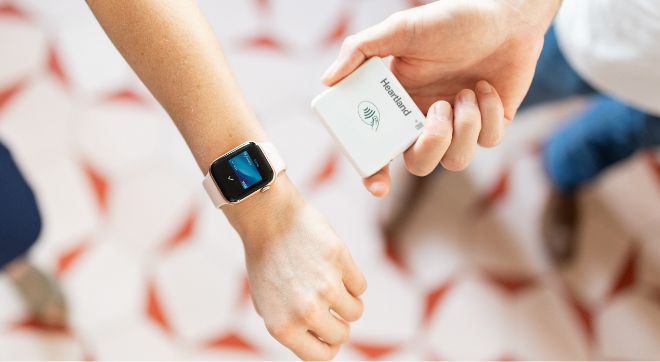 Person holds a smart watch next to a Heartland Mobile Pay terminal to make a contactless payment